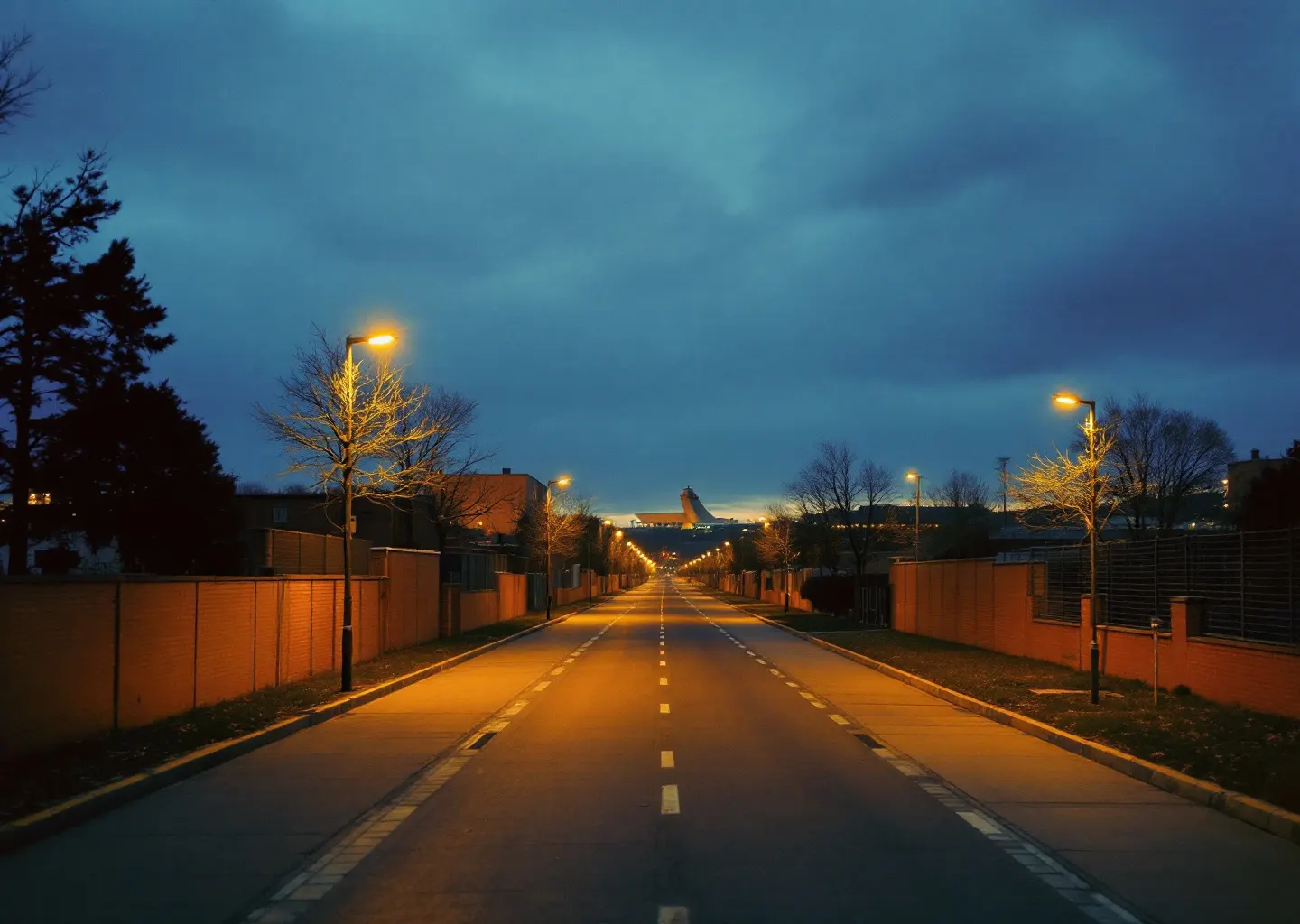 Long boulevard rectiligne au crépuscule longeant la clôture d'un site industriel, lampadaires allumés