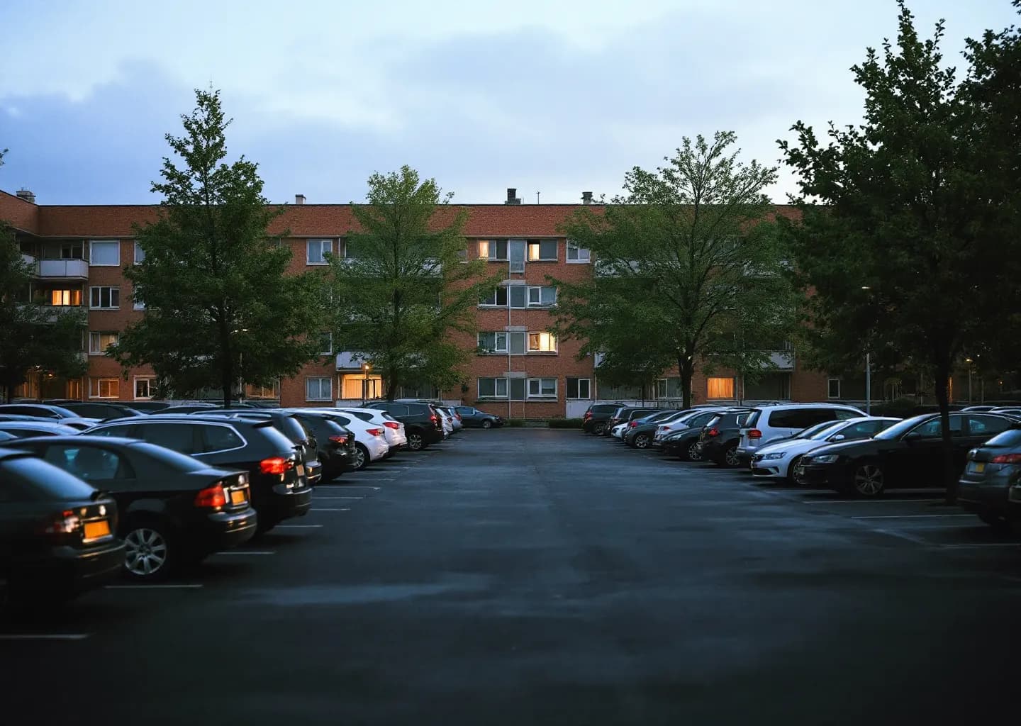 Parking d'un immeuble collectif aux premières lueurs, rangées de voitures et une seule fenêtre éclairée