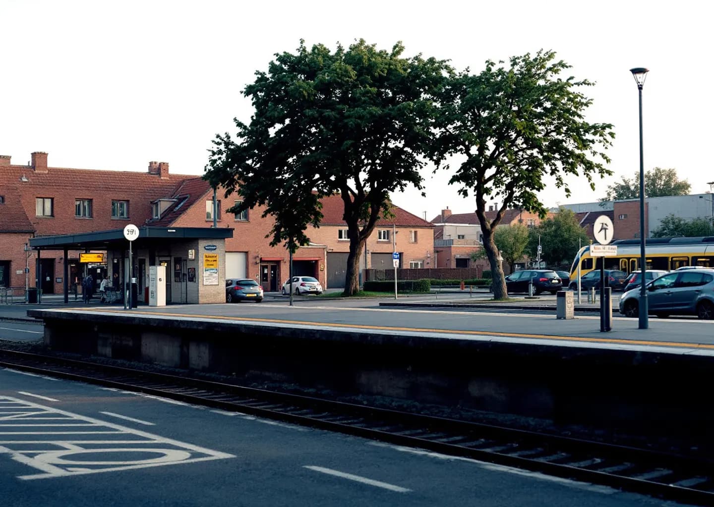 Petite gare de banlieue avec quai dégagé, parking-relais et entrepôts d'activité en arrière-plan